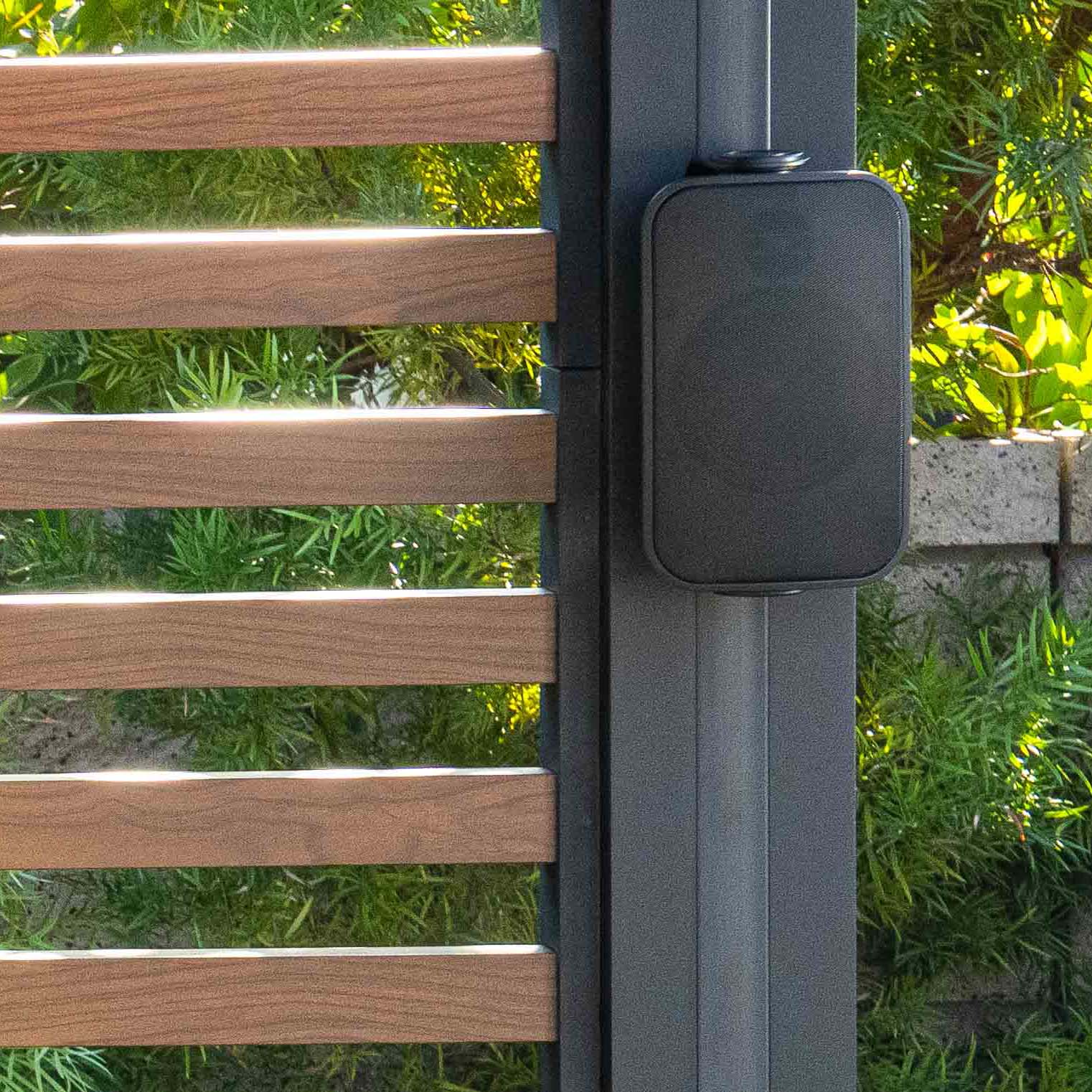 Black outdoor speaker mounted on a dark metal post beside a wood-slat fence with lush greenery.