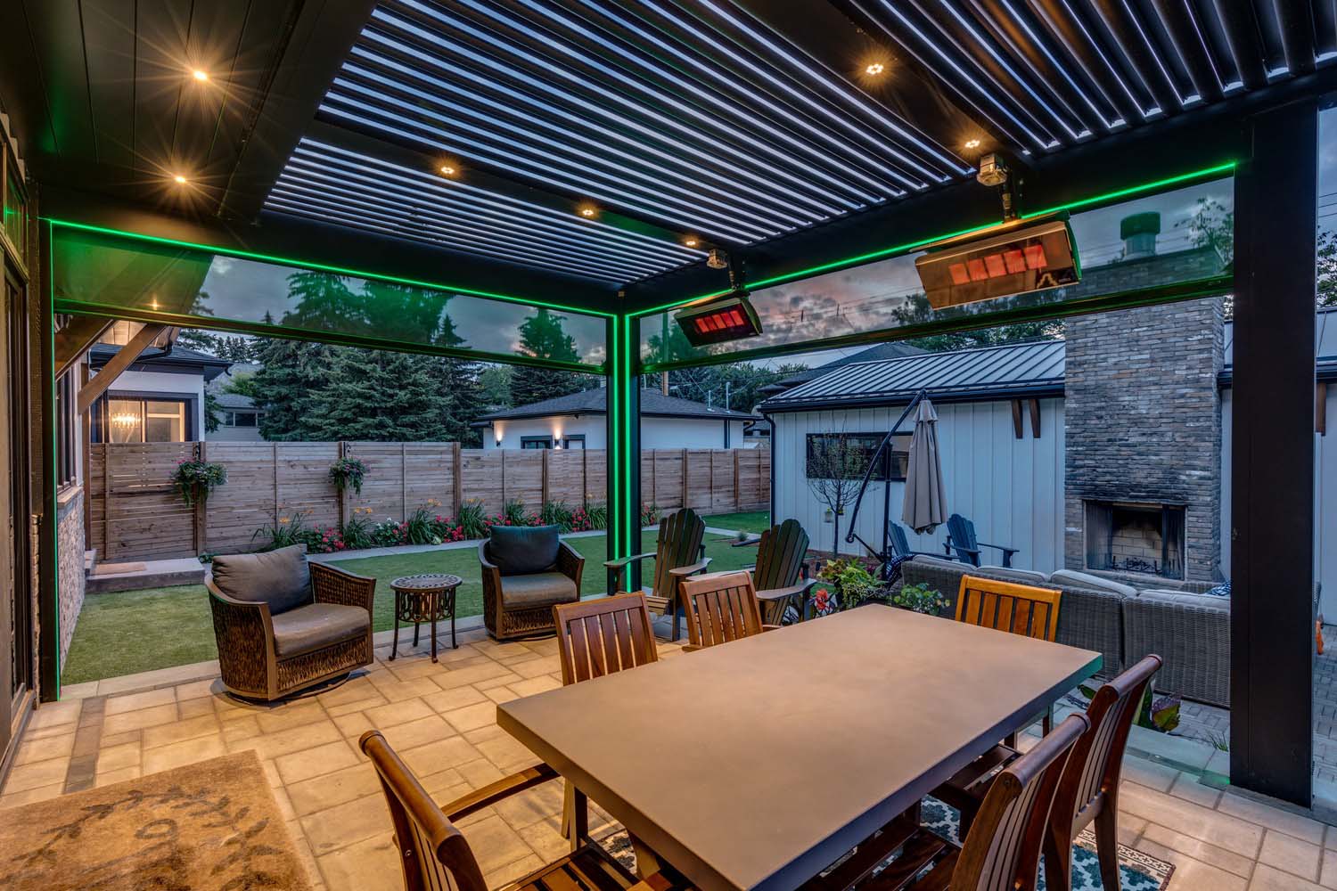 Covered outdoor patio with a slatted pergola roof, recessed lights, and green LED accent lighting, featuring a dining table, lounge seating, and a view of a landscaped backyard with a fireplace and additional seating.