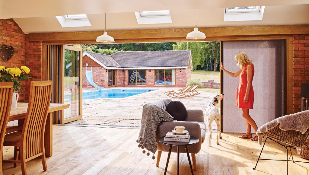 A woman and her Dalmatian dog stand inside a bright, open-concept living room featuring exposed brick walls and skylights. Large sliding glass doors open up to a spacious backyard patio and a luxury swimming pool.