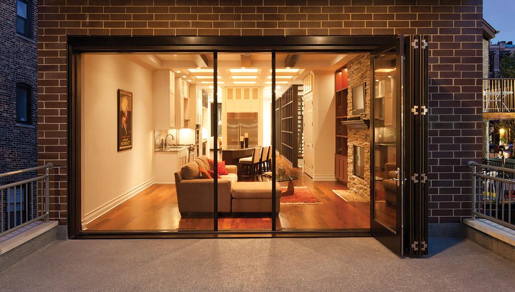 Modern open-concept condo with large bi-fold glass doors opening onto a spacious outdoor patio, showcasing a warm interior with a living room, stone fireplace, and kitchen.