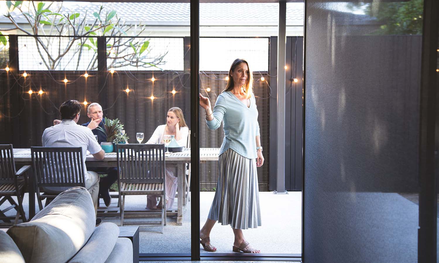 Woman sliding open a glass door to a patio where people dine under string lights.