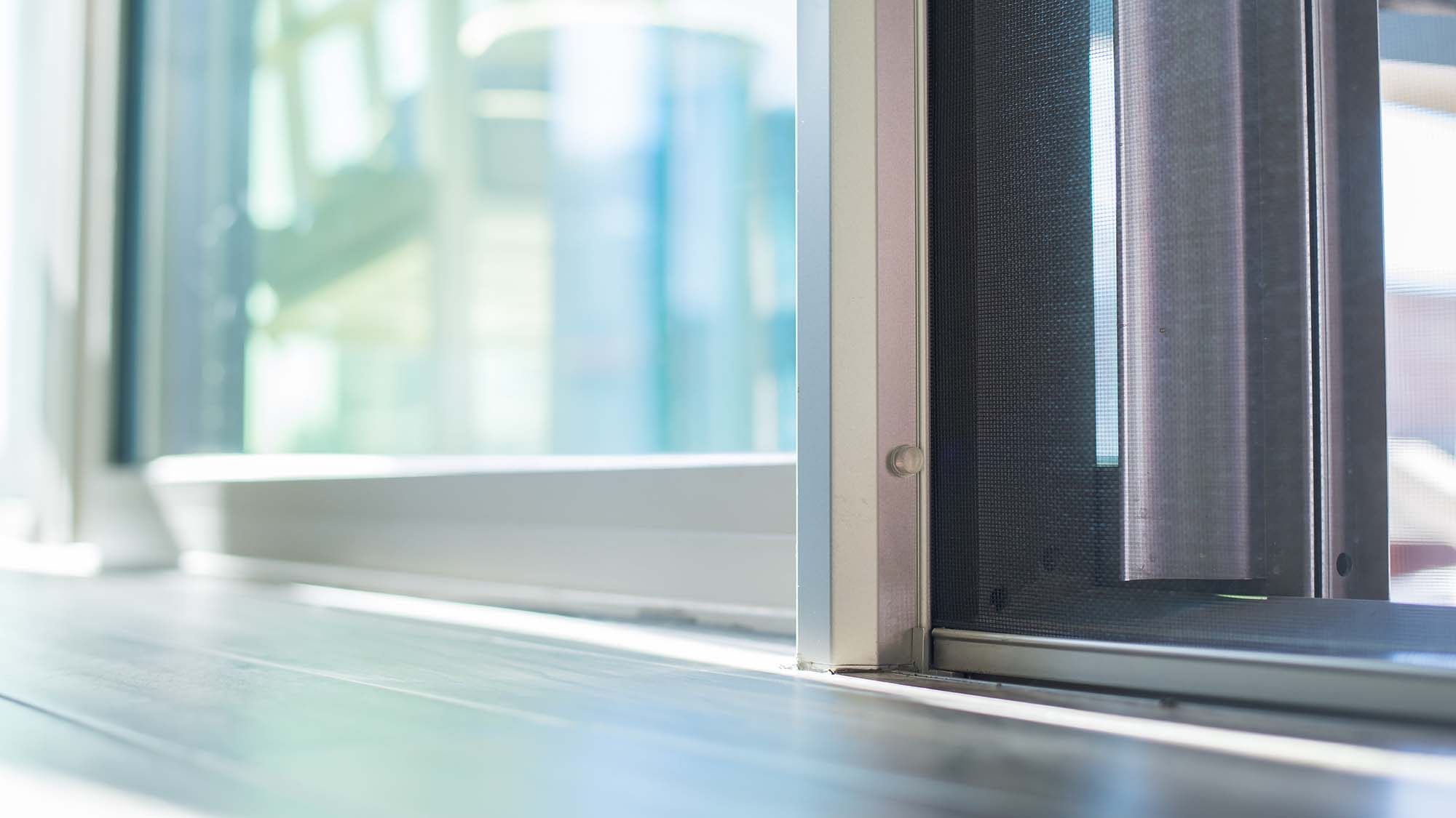 Close-up of a sunlit sliding glass door and screen at floor level, showing the metal track and blurred outdoor scene beyond.