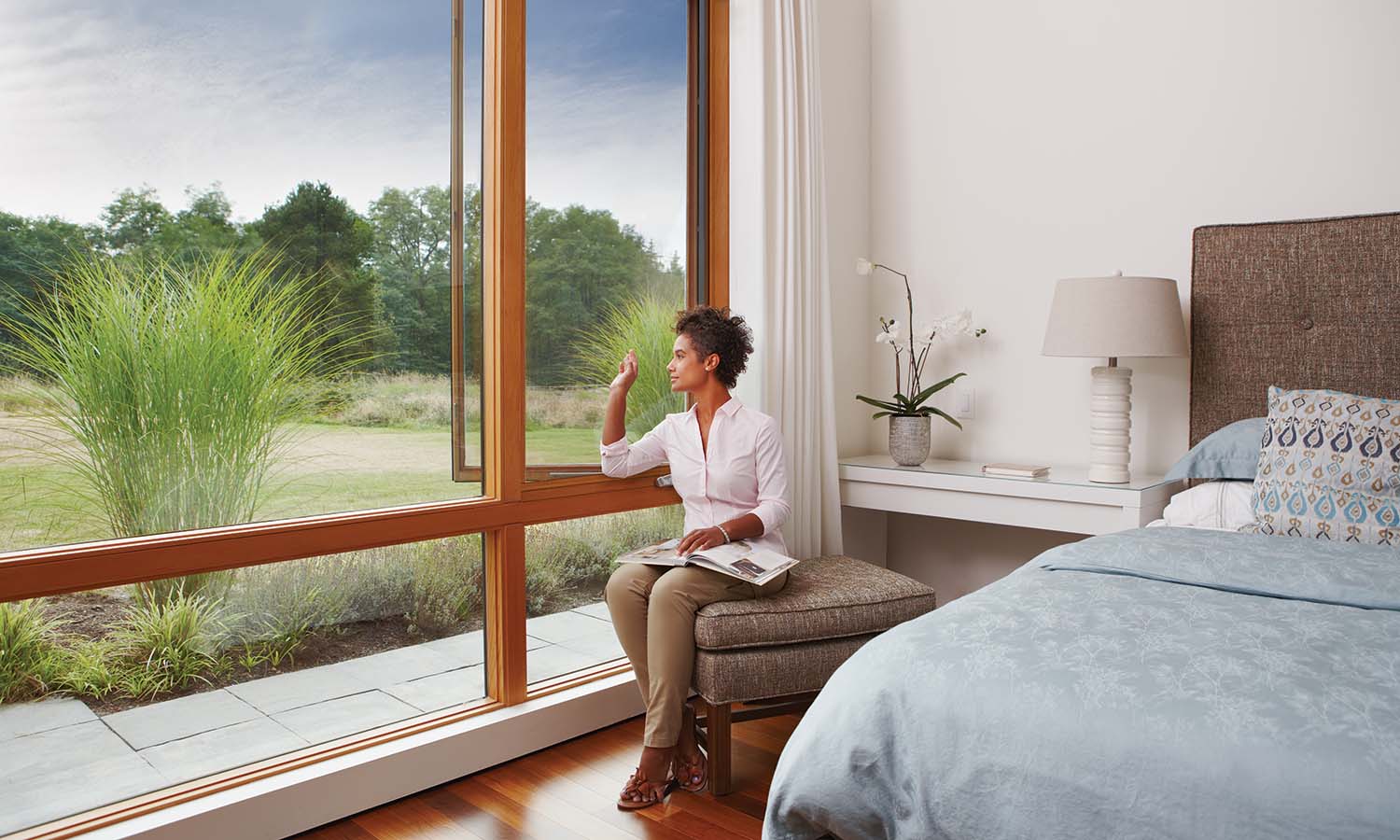 Woman seated by a large wood-framed bedroom window, looking outside across a grassy yard.