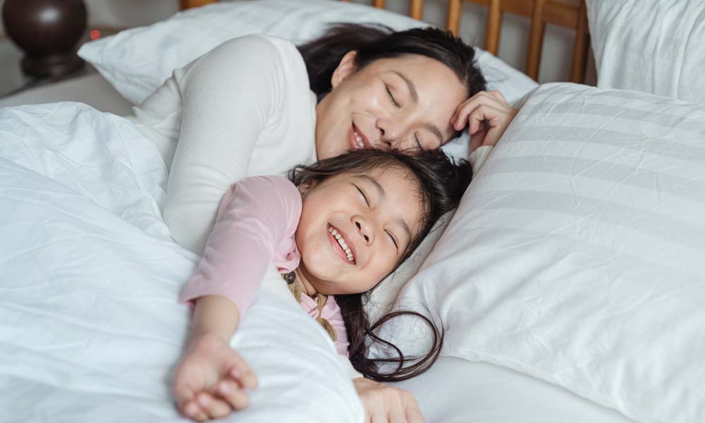 Smiling mother and young daughter cuddle under white bedding, lying face to face on a bed with their eyes closed and looking relaxed and happy.