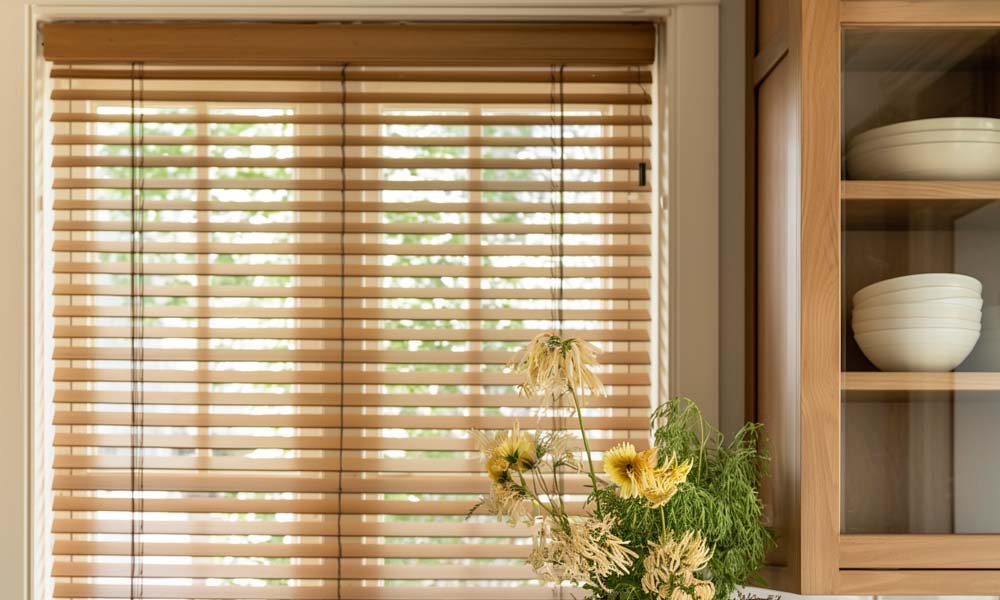 Close-up of a window with wooden blinds beside a kitchen cabinet and a vase of flowers.