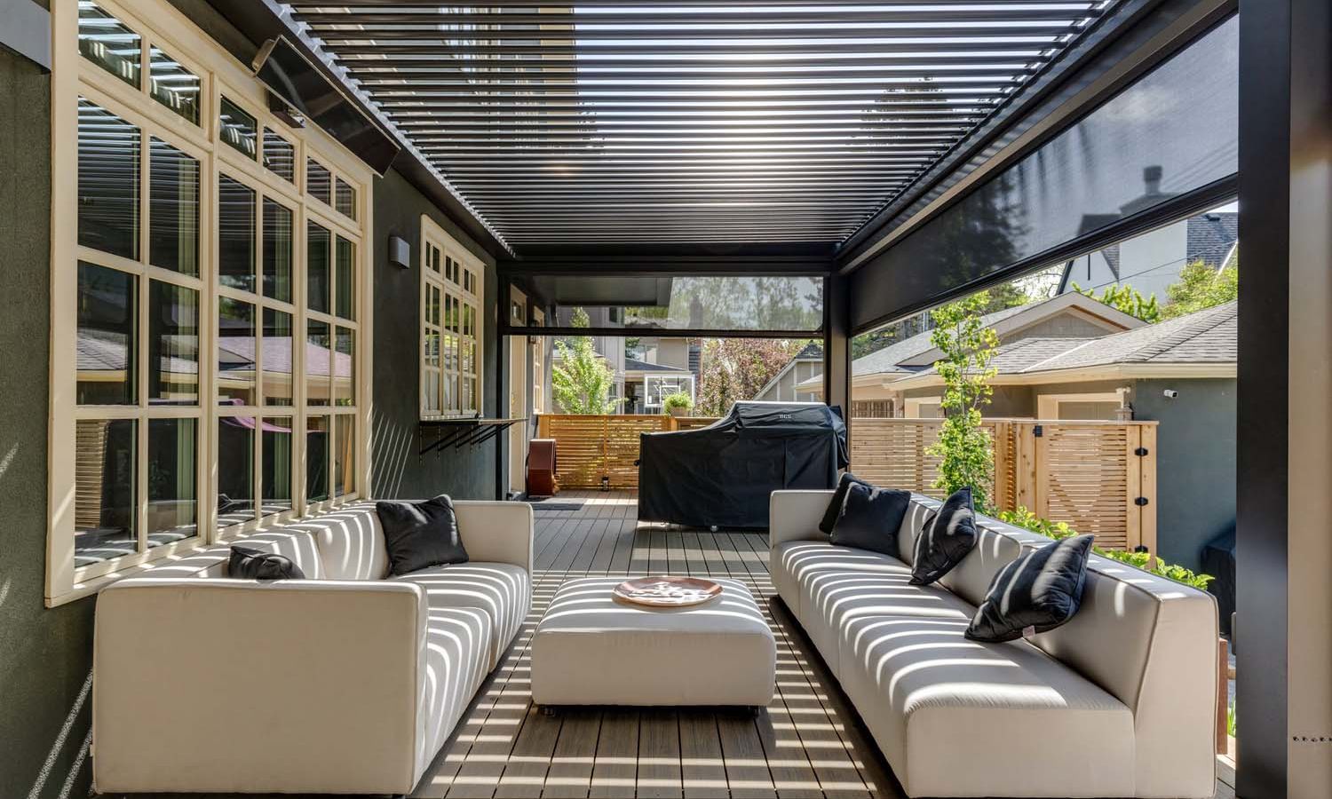 Modern backyard lounge under a louvered pergola with motorized drop screens; two long white sofas face a low ottoman while striped shadows from the louvers fall across the deck, with a covered grill and garden beyond.