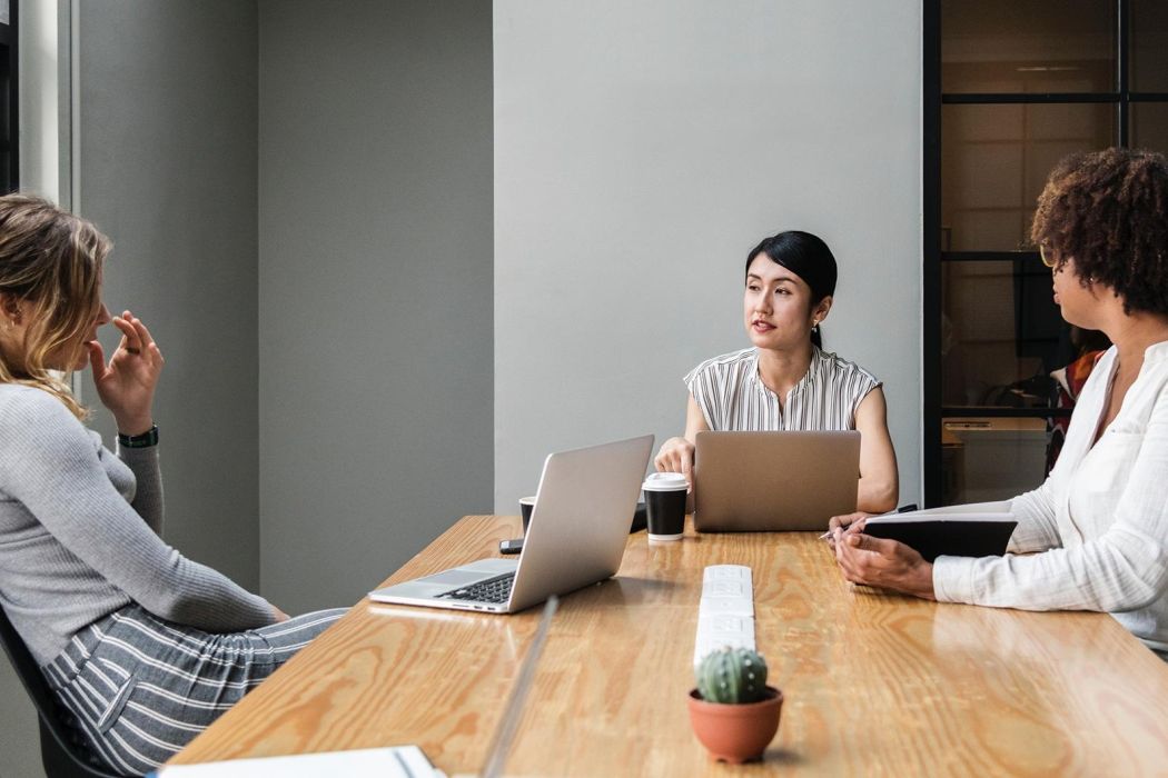 Three colleagues seated around a long wooden conference table in a modern office, laptops open and a coffee cup nearby, as the woman in the center speaks; a small potted cactus and built-in power strip run down the middle of the table.