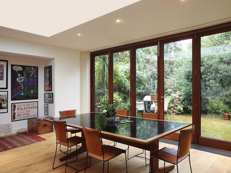 Modern dining room with a large table, wooden chairs, and wall art, featuring wide bi-fold glass doors that open to a lush green garden patio.