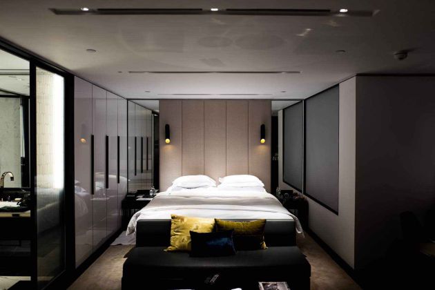 Contemporary bedroom with an upholstered headboard, mirrored closets, and wall sconces, featuring dark roller shades lowered to darken the room for restful sleep.