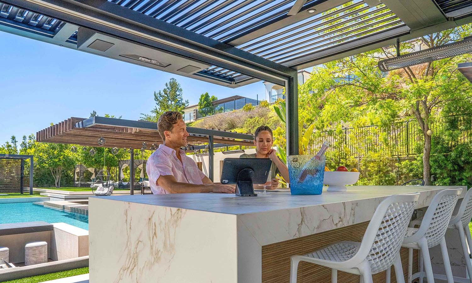 Couple sitting at a marble outdoor bar under a modern louvered pergola, looking at a tablet, with a swimming pool and landscaped backyard visible in the bright sunshine.