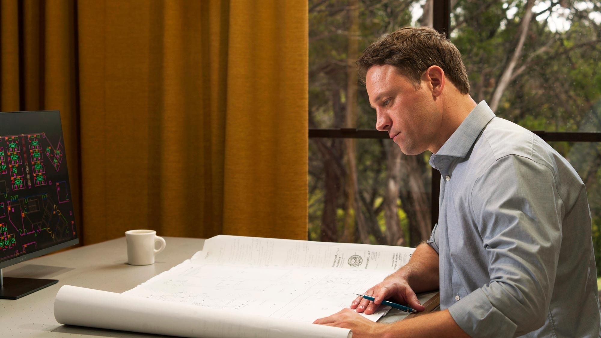 Man in a light blue shirt studies large architectural blueprints at a desk with a monitor displaying floor plans, a coffee mug, and yellow curtains framing a window that looks out onto trees.